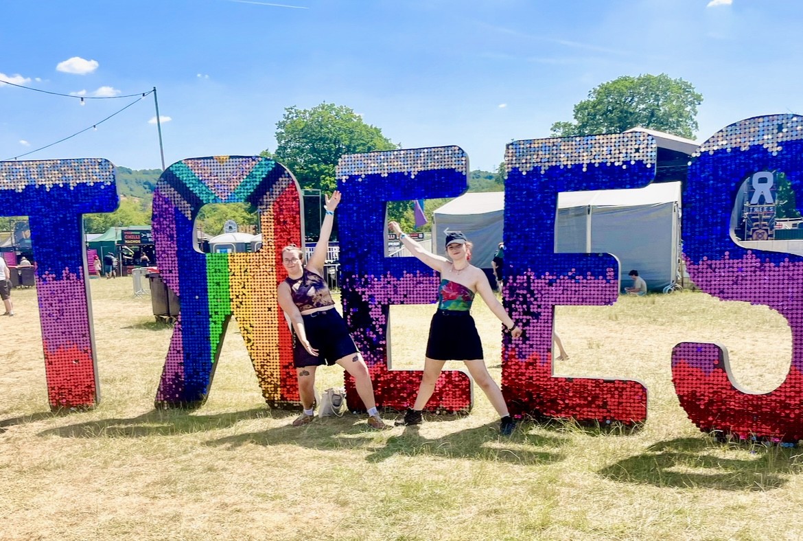 Festival For You team standing at the 2000trees Festival entrance with the Progress Pride flag, symbolising LGBTQ+ and trans inclusivity at UK summer music festivals 2025.